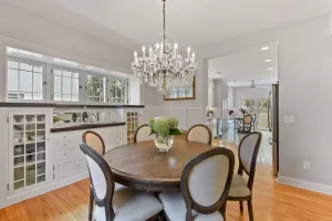 Elegant dining area with a round wooden table, six upholstered chairs, a glass vase with flowers, and a crystal chandelier. Built-in cabinets, large windows, and an open kitchen are visible in the background.