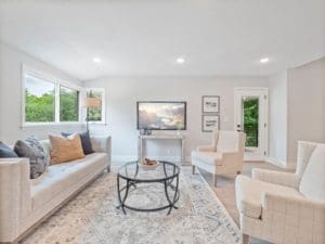 Bright living room with a beige sofa, two armchairs, a round glass coffee table on a patterned rug, and a TV on a stand. Large windows and a glass door let in natural light and views of greenery outside.