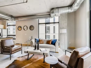 Modern living room with industrial decor, exposed ducts, large windows, a light sofa with colorful pillows, two brown leather chairs, a cowhide rug, and a concrete coffee table. Round mirrors hang on the wall.