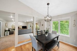 A modern open-concept dining and living area with light walls, wood flooring, a black dining table with gray chairs, large windows overlooking greenery, and a cozy living space with gray sofas and a fireplace.
