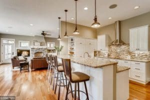 Modern open-concept kitchen and living area with hardwood floors, white cabinets, granite countertops, pendant lights, and a stone fireplace. Three barstools line the island near a cozy seating area.