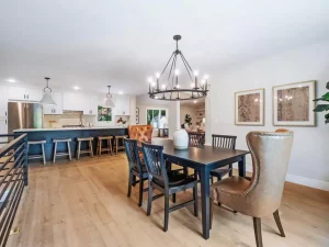 Open dining area with a black table, assorted chairs, modern chandelier, and adjacent kitchen with barstools, white cabinets, stainless steel appliances, and pendant lights. Light wood flooring throughout.