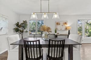 A modern dining room with a dark wood table, four white cushioned chairs, two dark wooden chairs, a large glass vase with dried palm leaves, abstract artwork, a potted plant, and large windows letting in natural light.