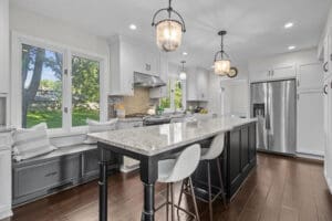 Bright, modern kitchen with white cabinets, stainless steel appliances, a large island with bar stools, pendant lights, and a window seat overlooking a green backyard. Dark wood floors contrast with the light décor.