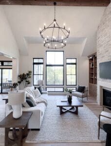 Bright living room with large windows, a white sofa, neutral decor, a wooden coffee table, a modern chandelier, a stone fireplace, built-in shelves, indoor plants, and light wood flooring.