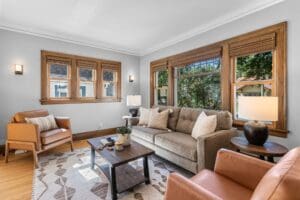 Bright living room with large windows, a gray sofa, two tan armchairs, wooden coffee table, patterned rug, and neutral decor. Natural light fills the space, highlighting wood trim and greenery outside.