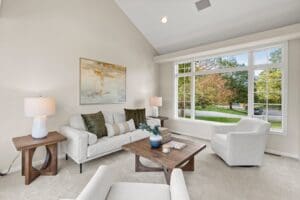 A bright, modern living room with neutral tones features a light sofa, two armchairs, wooden tables, a large window letting in natural light, and a painting on the wall above the couch.