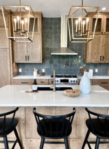 Modern kitchen with wooden cabinets, a white island with a gold faucet, three black chairs, geometric green tile backsplash, stainless steel stove, and two large gold pendant lights. Decor includes a vase, bottle, and wooden bowl.