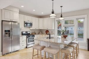A bright, modern kitchen with white cabinets, stainless steel appliances, granite island with four beige stools, pendant lights, and large windows letting in natural light. A plant sits on the island.