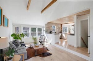 Bright, modern living room with light wood floors, exposed ceiling beams, a gray sofa, plants, and a cowhide rug, opening into a white kitchen with natural light and wooden accents.