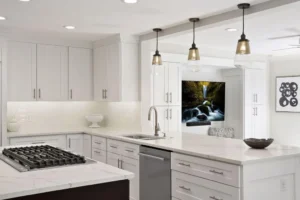 Modern white kitchen with marble countertops, stainless steel appliances, gas cooktop on the island, pendant lights, and a window-like opening revealing a living area with a waterfall image on the TV.