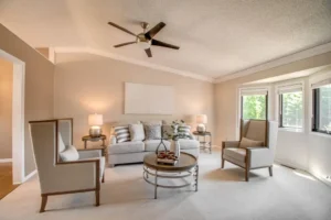 A bright, neutral-toned living room with a gray sofa, two armchairs, a round coffee table, table lamps, wall art, and large windows letting in natural light. A ceiling fan hangs from the textured ceiling.