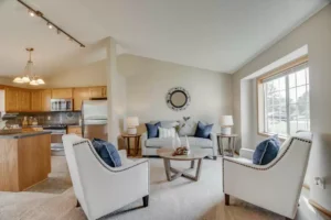 A bright living room with light walls, a sofa, two armchairs with blue pillows, a round wooden coffee table, two side tables with lamps, and a large window. The kitchen with wooden cabinets is visible in the background.