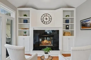 A bright living room with a white fireplace, built-in shelves, a large clock above the mantel, two white armchairs, a small round table with a potted plant, and framed art on the wall.