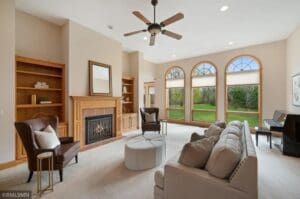 Bright living room with beige walls, large arched windows, a ceiling fan, a fireplace with a wooden mantel, built-in shelves, a gray sofa, armchairs, and a piano near the windows overlooking greenery.