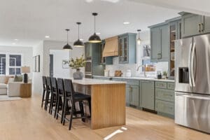 Modern kitchen with sage green cabinets, stainless steel appliances, a large island with wooden base and white countertop, black barstools, pendant lights, and light wood flooring. A living area is visible in the background.