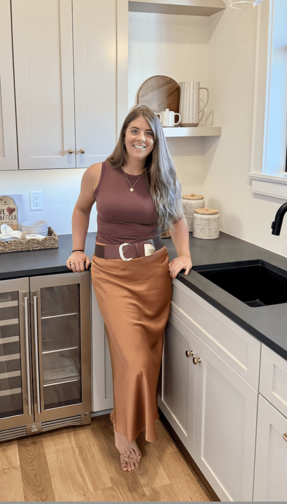A woman with long brown hair, wearing a sleeveless brown top and a shiny bronze maxi skirt with a wide belt, stands barefoot in a modern kitchen, smiling and leaning against a counter near a sink and wine fridge.