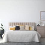 Modern bedroom with a large bed featuring a beige headboard, white and yellow pillows, a textured white bedspread, a light wood dresser, green potted plants, and a framed picture on a plain white wall.