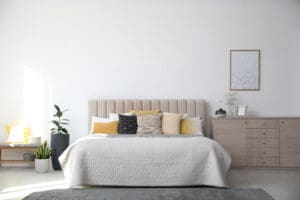 Modern bedroom with a large bed featuring a beige headboard, white and yellow pillows, a textured white bedspread, a light wood dresser, green potted plants, and a framed picture on a plain white wall.