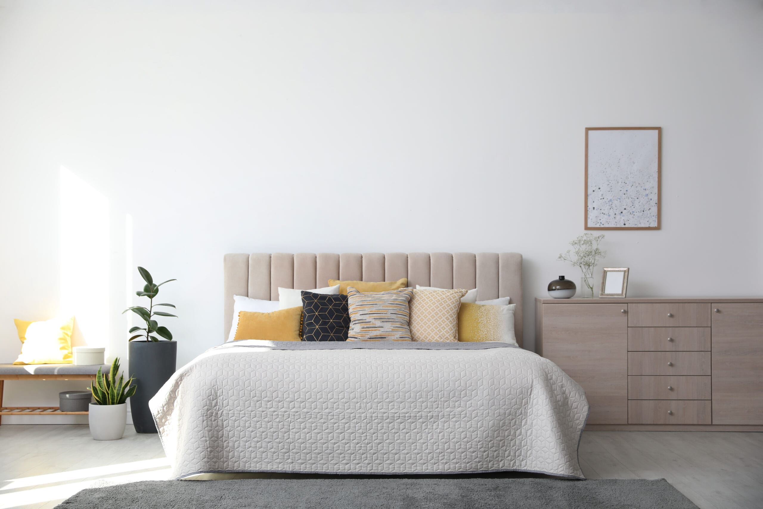 Modern bedroom with a large bed featuring a beige headboard, white and yellow pillows, a textured white bedspread, a light wood dresser, green potted plants, and a framed picture on a plain white wall.