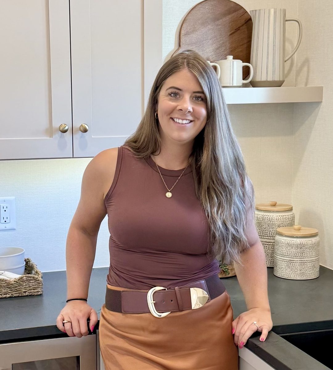 A woman with long brown hair, wearing a sleeveless maroon top and a brown skirt with a large belt, stands and smiles in a modern kitchen with neutral-toned decor and shelves holding dishes.